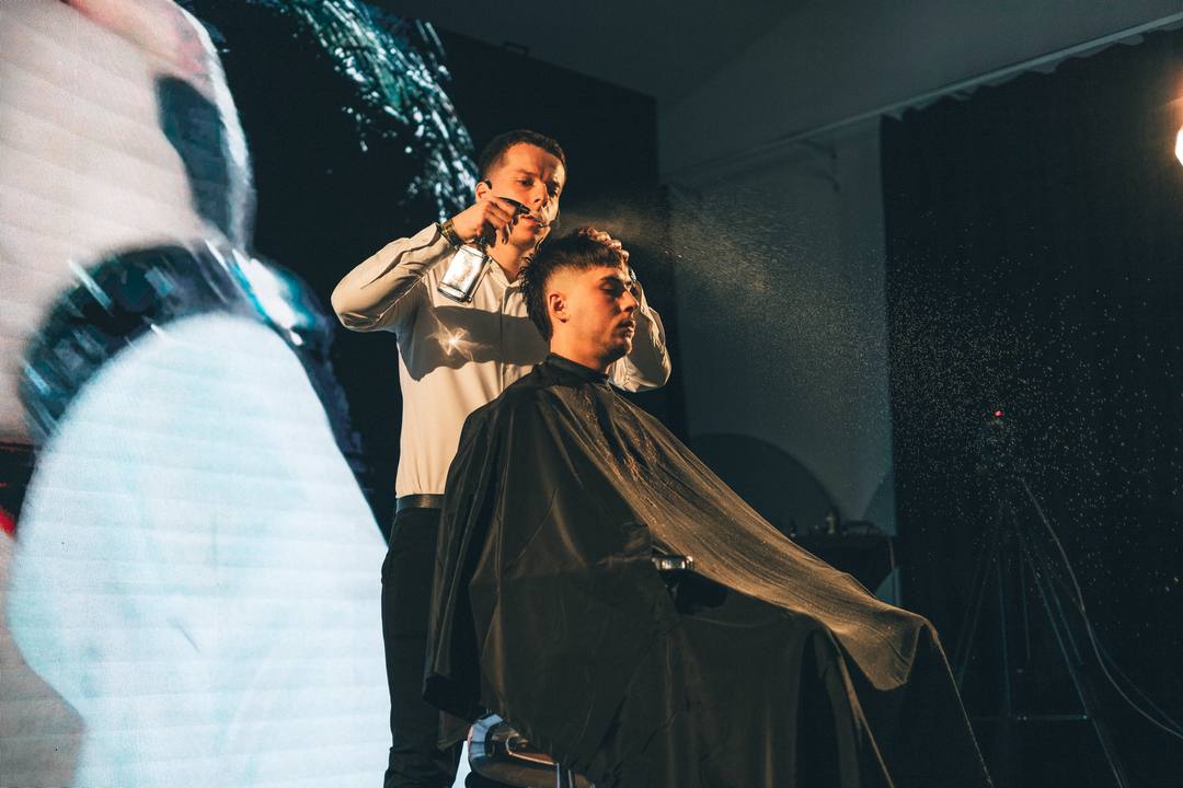 Vilis Sliacky upravuje vlasy modela na pódiu, vedľa neho veľkoplošná obrazovka