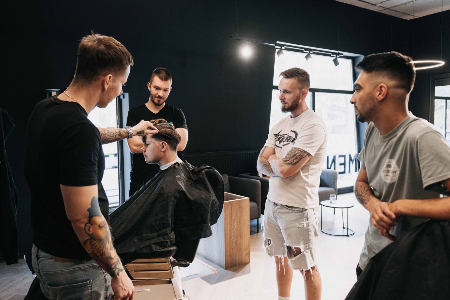 Lektor Vilis Sliacky predvádza strih na modelovi v barberskom kresle, traja študenti stoja okolo a sledujú ho